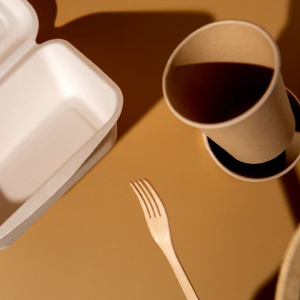 Paper coffee cup with dark coffee, white takeout container, and a wooden fork on a brown table top.