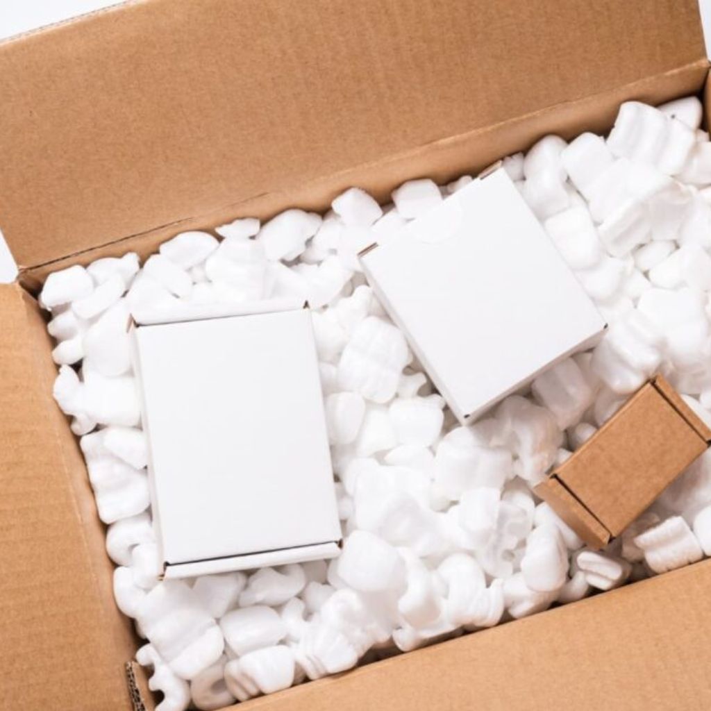 Open cardboard shipping box filled with white packing peanuts and several white and brown cardboard boxes inside.