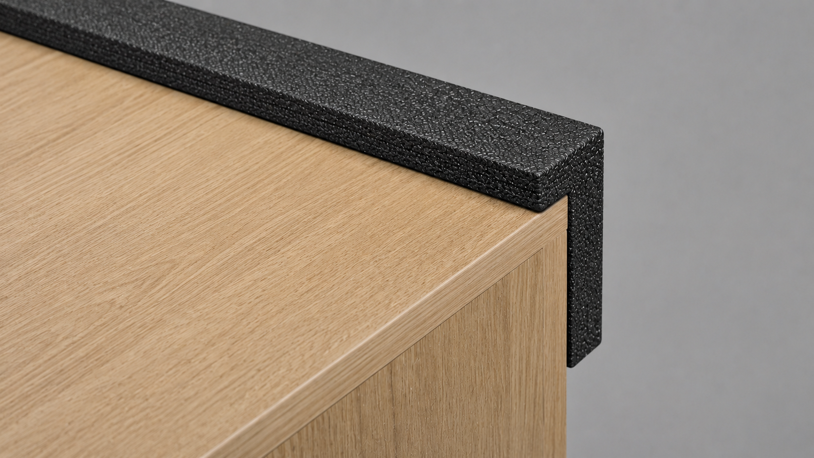 Close-up of a black speckled edge guard along the top corner of a light wood desk. Used to protect the edge from damage.